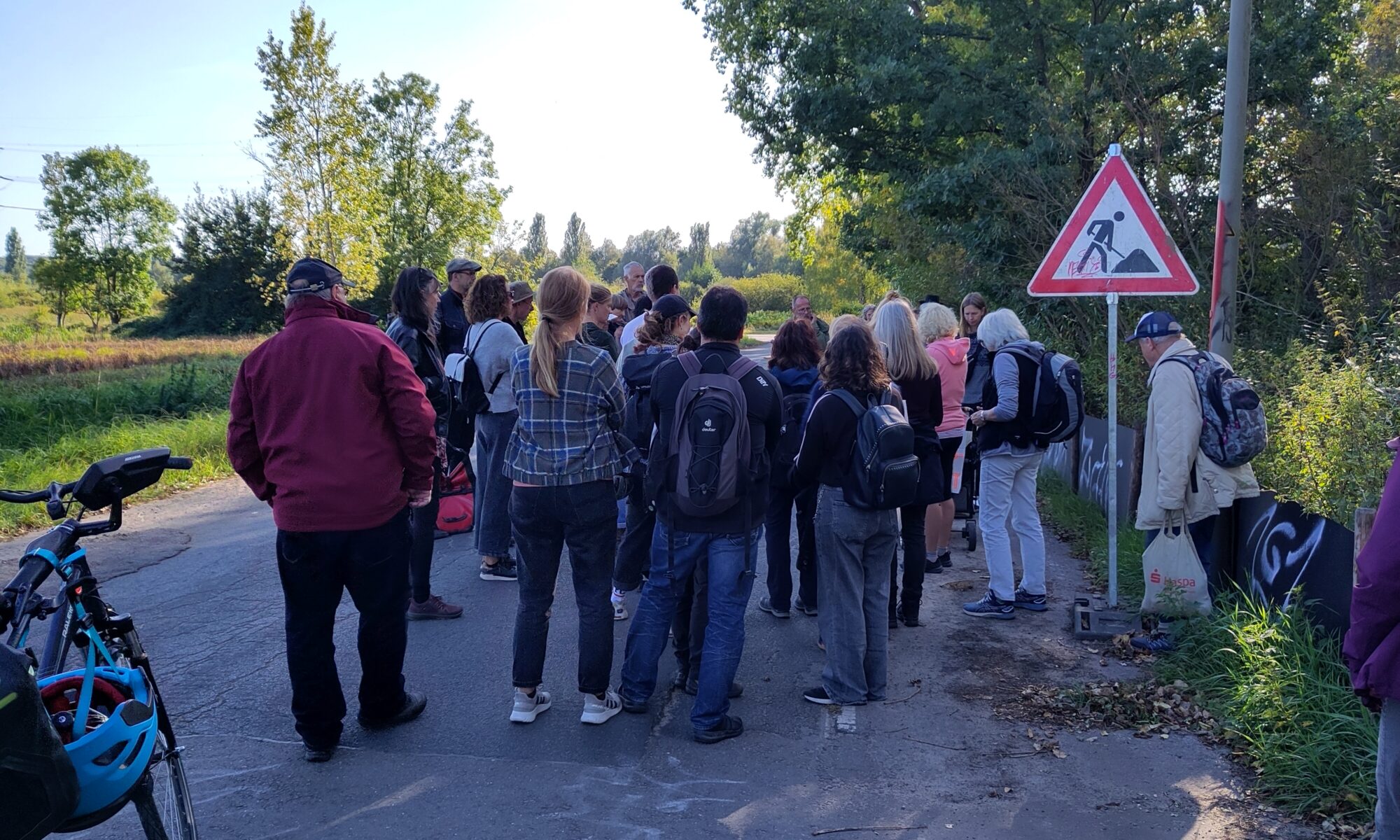 Eine Gruppe von Mesnchen staht mit dem Rücken zur Betrachter*in auf einer kleinen Straße am Waldrand und hört einer Person zu. Rechst neben der Gruppe ein Baustellenschild. Die Gruppe steht im Schatten, im Hintergrund baluer, sonniger Himmel.