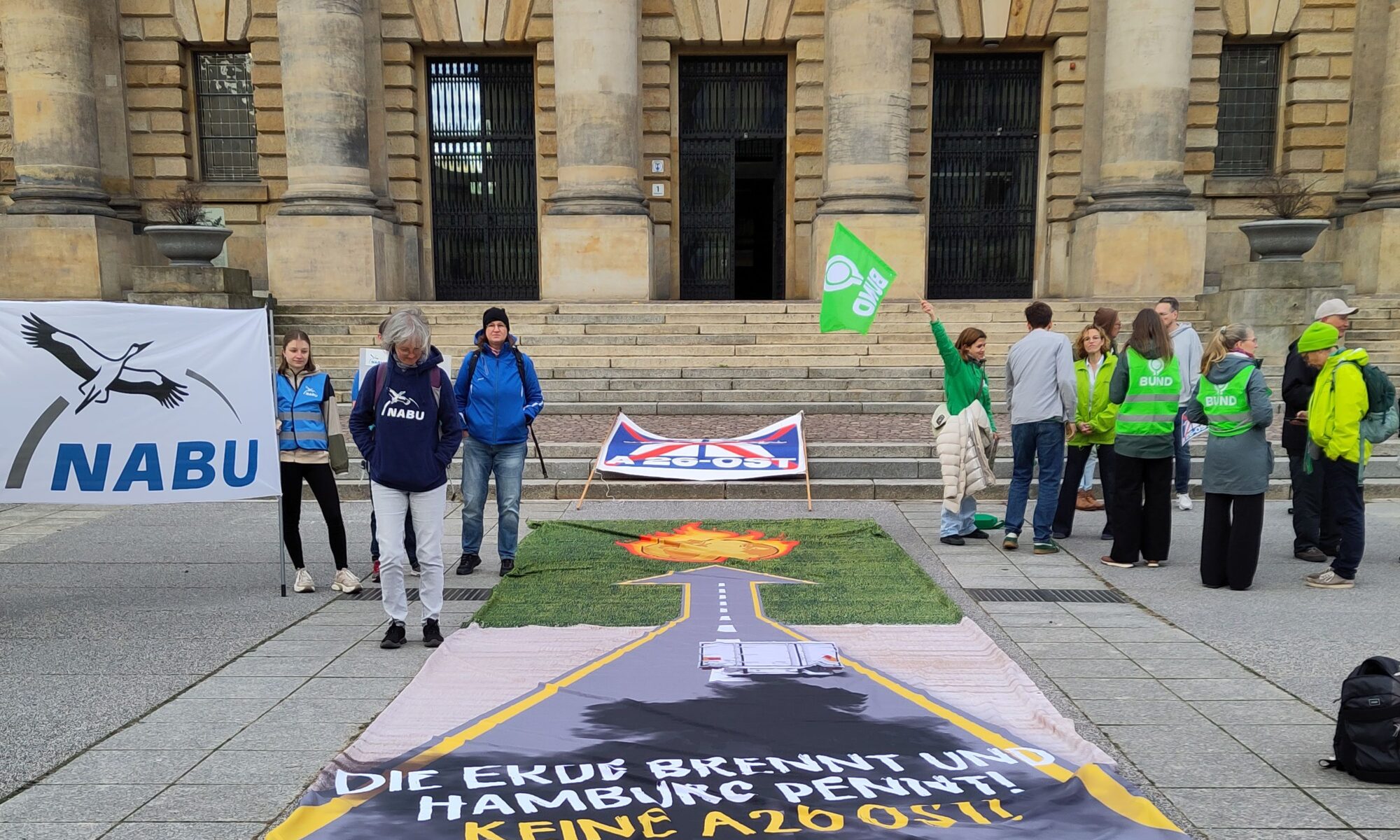 Vor den steinernen Treppen des riesigen, altehrwürdigen Gebäudes des Bundesverwaltungsgerichtes in Leipzig liegt das lange Banner: eine Autobahn führt in die Ferne, endet mit einem Pfeil auf grüner Wiese, der Pfeil weist auf ein loderndes Feuer. Text unten: Die Erde brennt und Hamburg pennt. Keine A 26 ost. Auf der Treppe das Banner mit dem Verkehrsschild Autobahn (blau), dass mit rot durchkreuzt ist, unten steht: A26 Ost. Rechts stehen Leute mit blauen und weißen Kleidungsstücken und einer weißen Fahne mit Aufschrift NABU, rechts vom langen Banner Leute mit grünen Kelidungsstücken und einer kleinen grünen Fahne mi dem Logo des BUND.